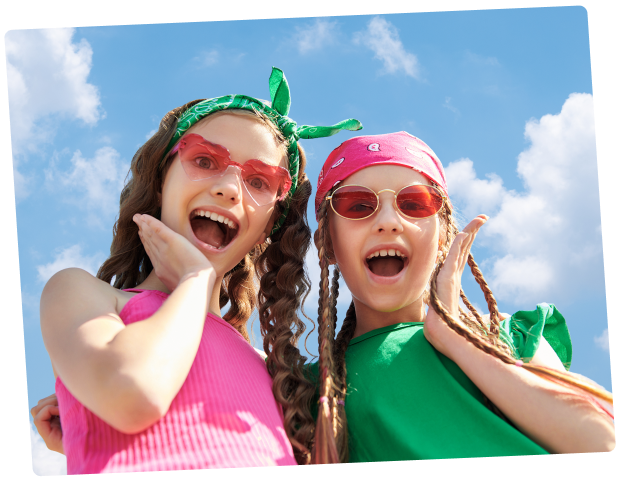 Two young girls outdoors under a blue sky with clouds, wearing bright pink and green outfits with sunglasses, headbands, and braided hair.